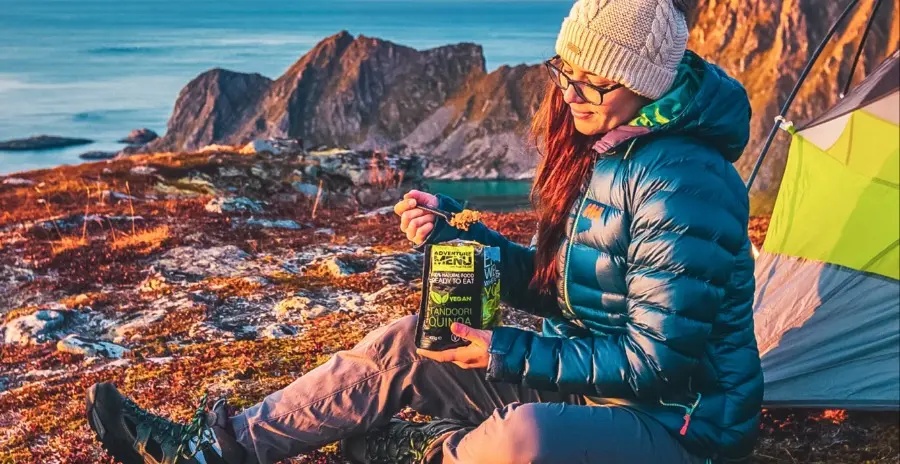 žena sedí před stanem u vody a jí hotovou mre snídani přímo ze sáčku