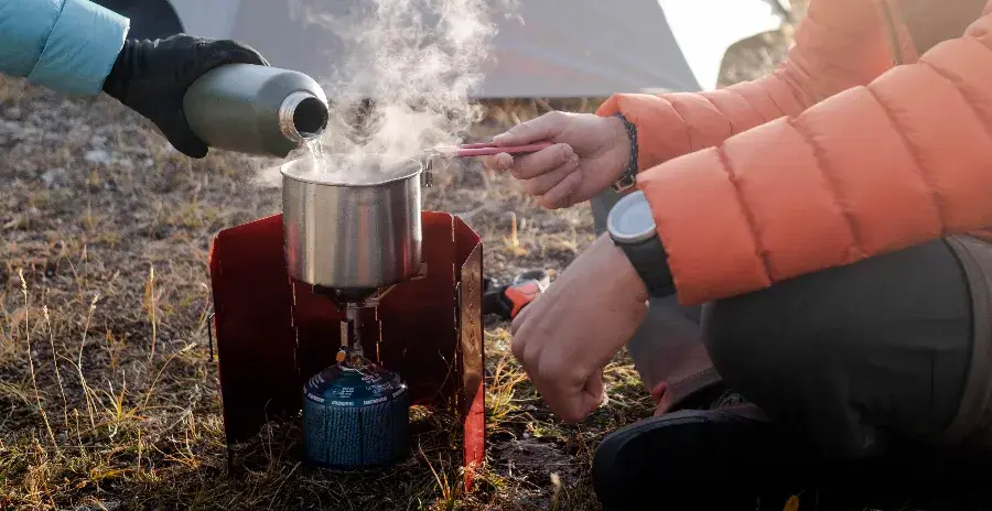 muž vaří před stanem jídlo na vařiči a kolem vařiče má zástěnu proti větru